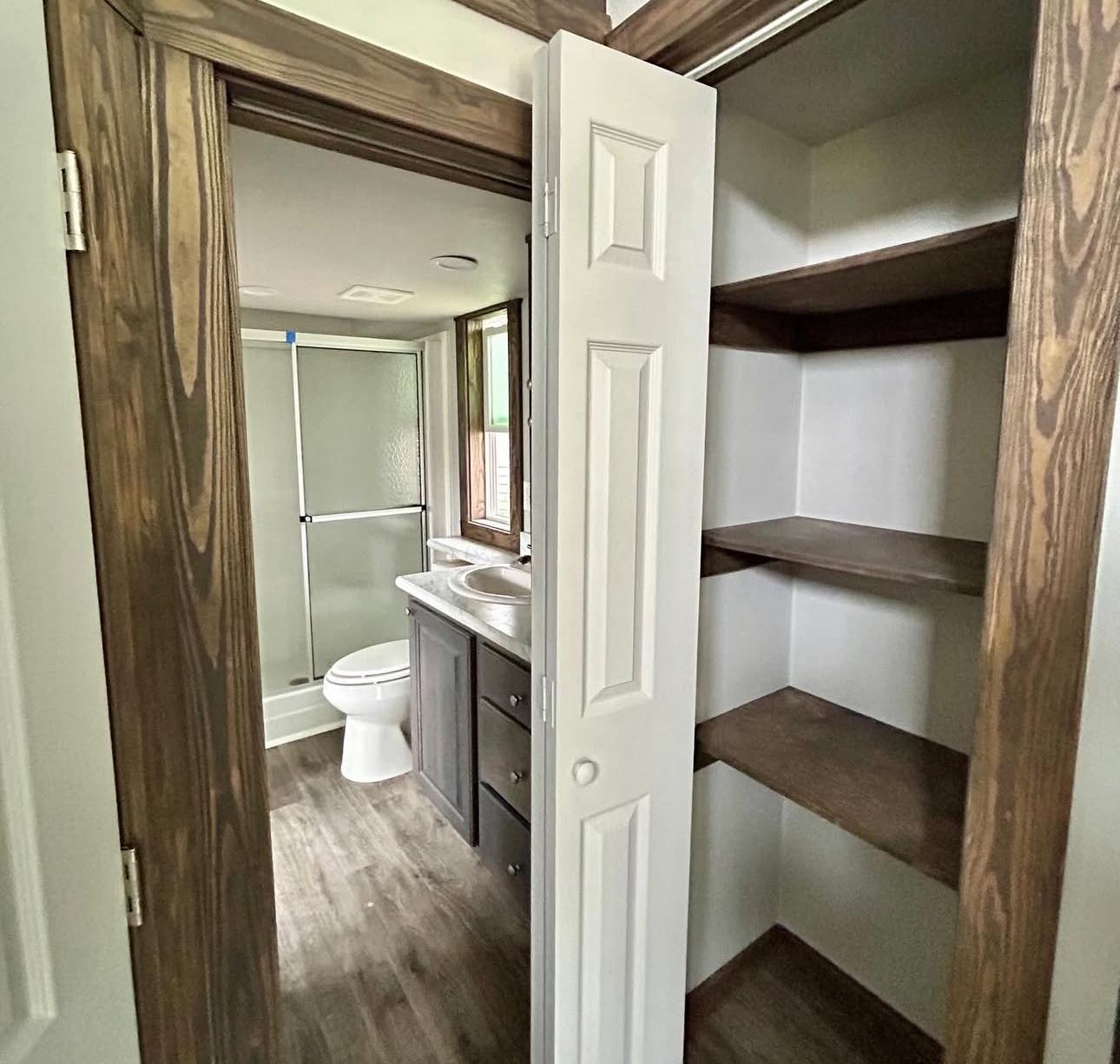 Compact bathroom with a glass shower door, toilet, and sink below a window. Adjacent open closet with wooden shelves, creating a cozy, practical vibe.