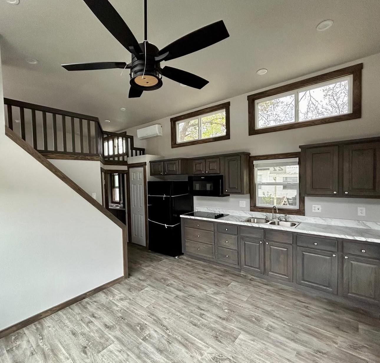 Modern kitchen with gray cabinets, marble countertops, black fridge, and microwave. A wooden stairway leads to a loft, and the floor is gray wood.