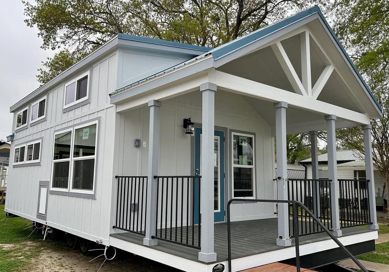 Modern tiny house with a gabled roof and blue trim, featuring a covered porch and black railings. Large windows add to the bright and airy feel.