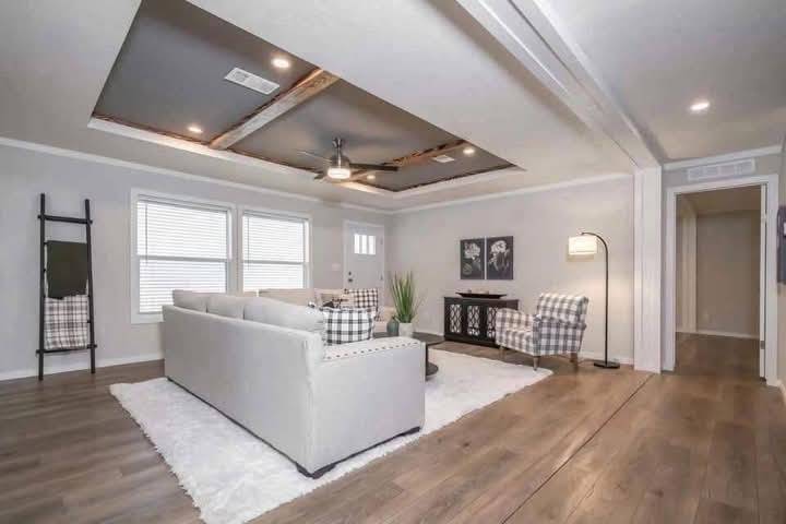 A cozy living room with a modern farmhouse style. It features a gray sectional sofa, plaid armchairs, a white rug, wooden floors, and wall art. The ceiling has wood beams, creating a warm and inviting atmosphere.
