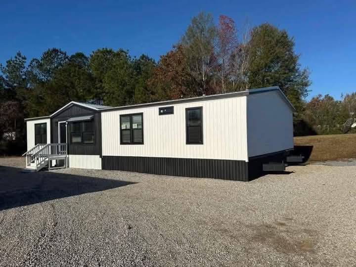 A white modular home with black trim sits on a gravel lot. It has a small front porch with stairs, backed by a line of tall trees on a clear day.