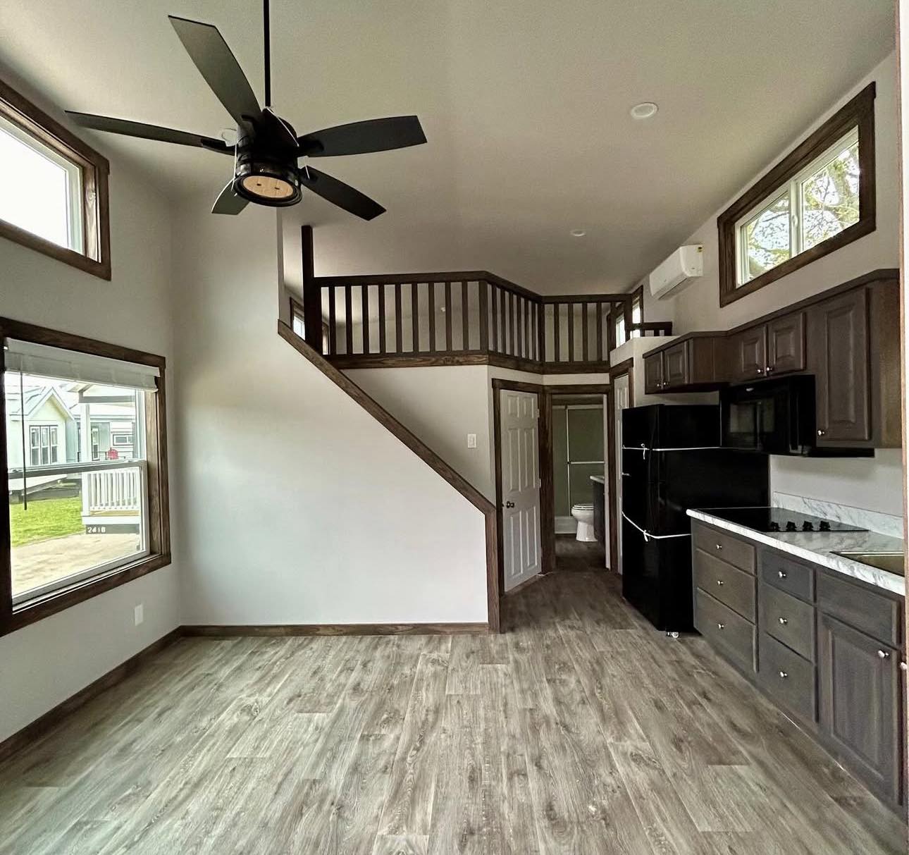 Spacious tiny home interior with laminate flooring, modern kitchen cabinets, black appliances, large windows, a ceiling fan, and a wooden loft above.