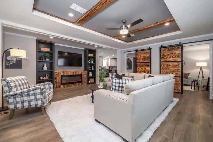 Stylish living room with modern gray and wood accents. Cozy plaid chair, beige sofa, wall-mounted TV, black shelves, sliding barn doors, and ceiling fan.
