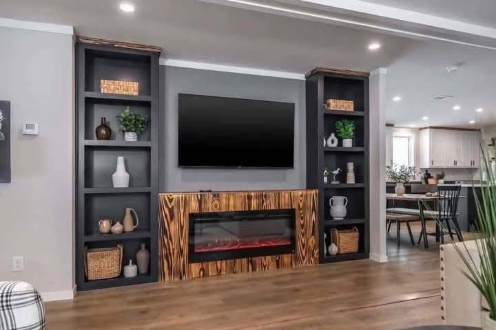 Modern living room with a wall-mounted TV above a wooden fireplace. Black shelves with plants and decor flank the fireplace. Open kitchen visible in the background.