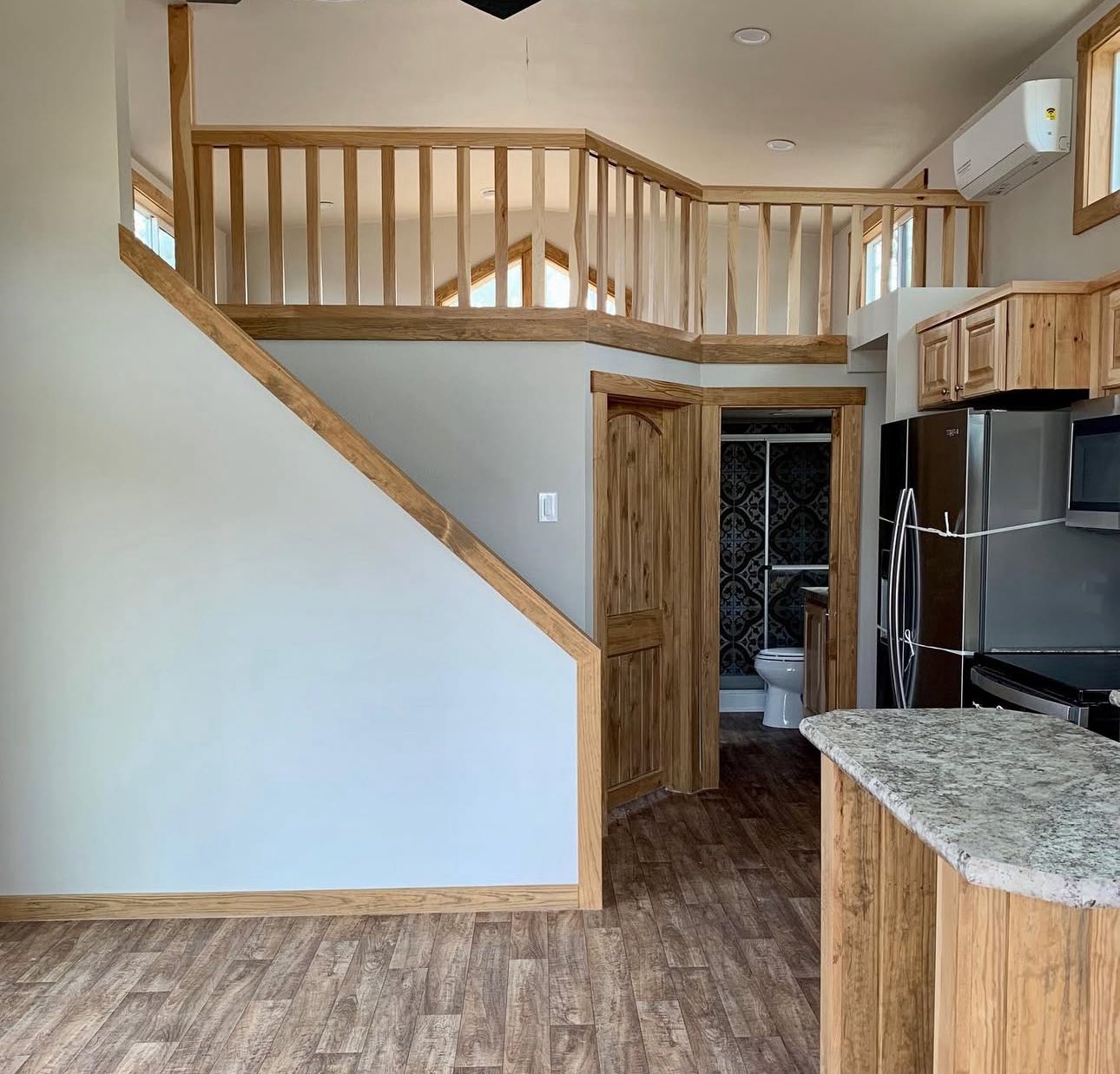 Cozy interior of a compact home featuring wooden accents. A staircase leads to a loft with railing. The kitchen has stainless steel appliances and granite countertops.