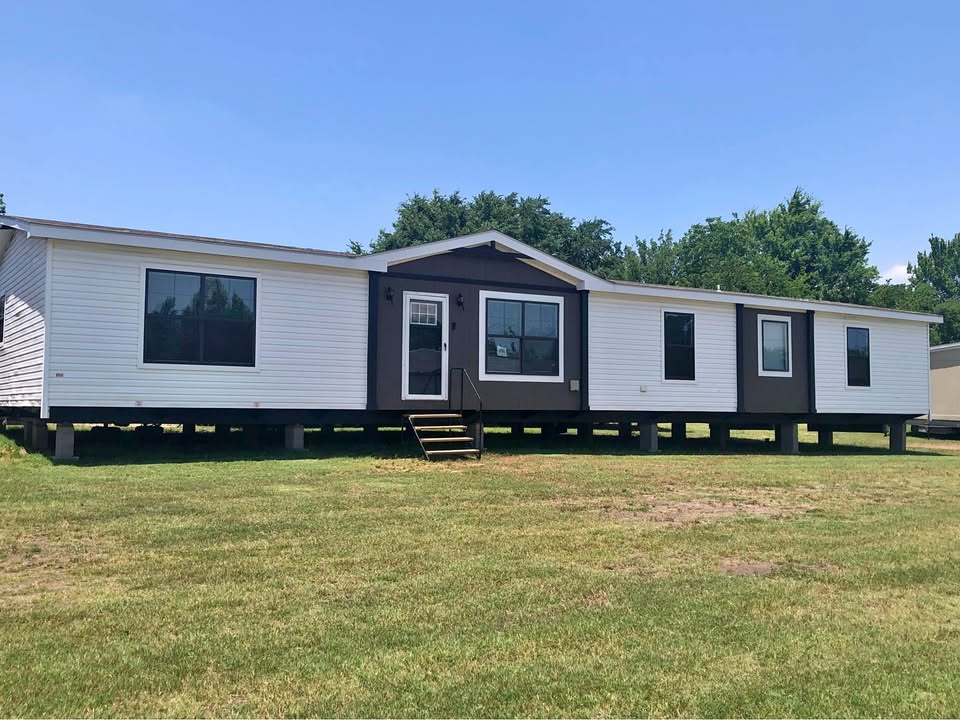 A manufactured home with white and dark trim sits elevated on a grassy lawn. The sky is clear, with trees in the background, creating a serene setting.