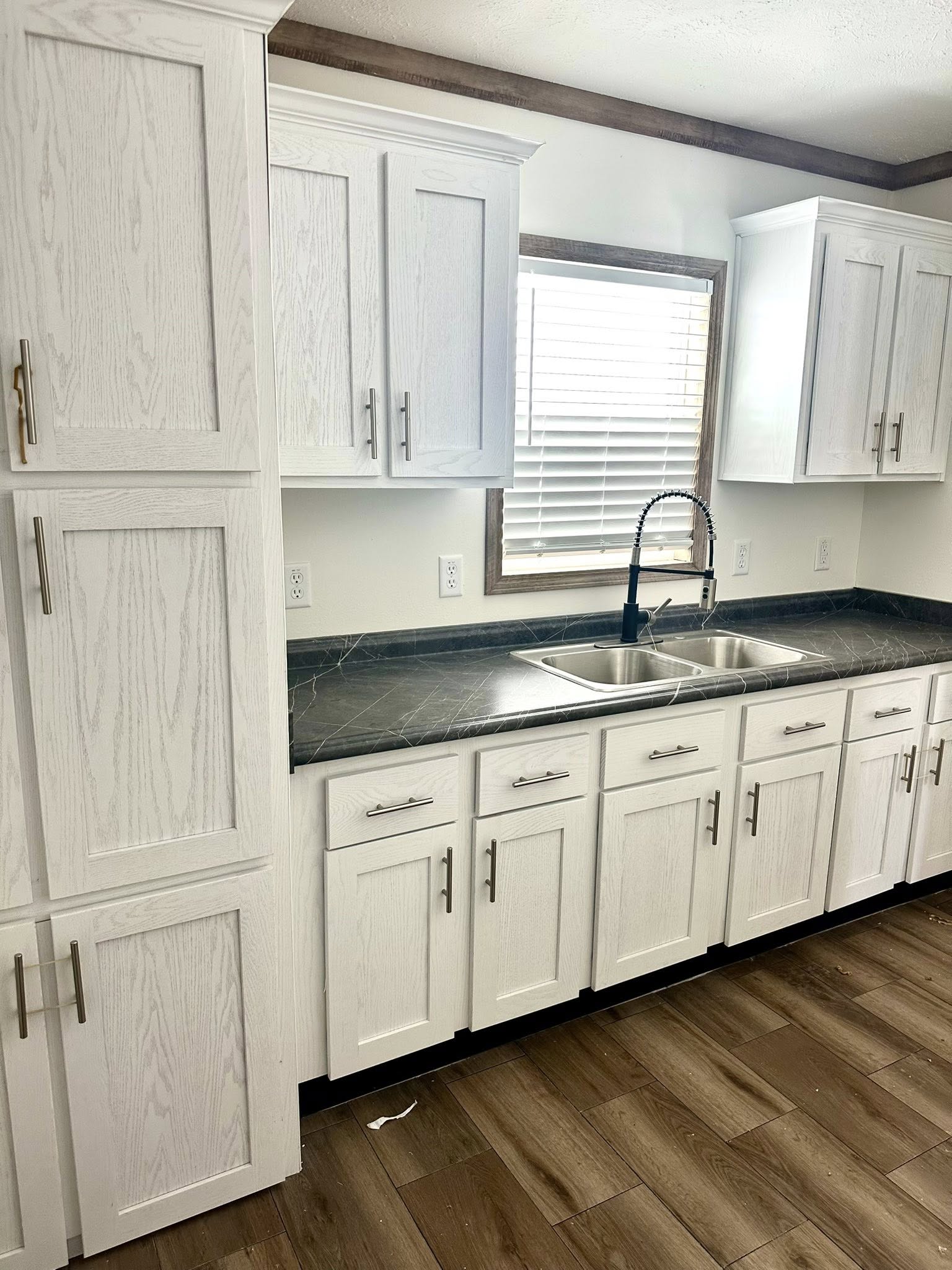 Modern kitchen with white cabinets, dark marble countertop, and stainless steel dual sink. A window with blinds allows light in, highlighting wood flooring.