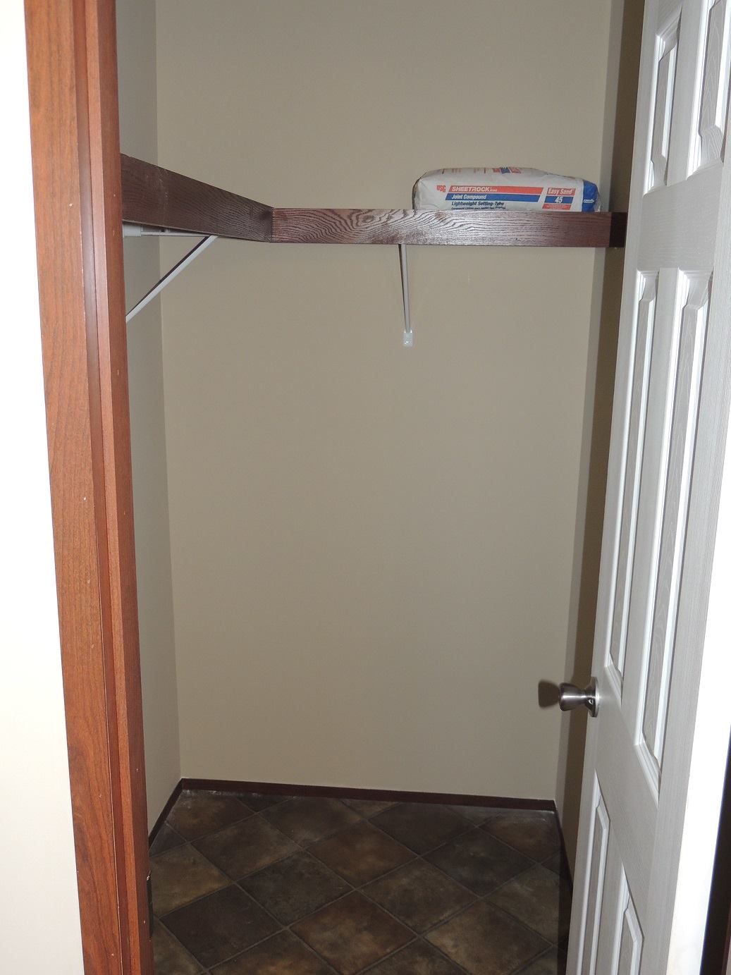 An empty closet with beige walls, dark wooden shelf, and tiled floor. A single packaged item sits on the shelf, with the door slightly ajar.