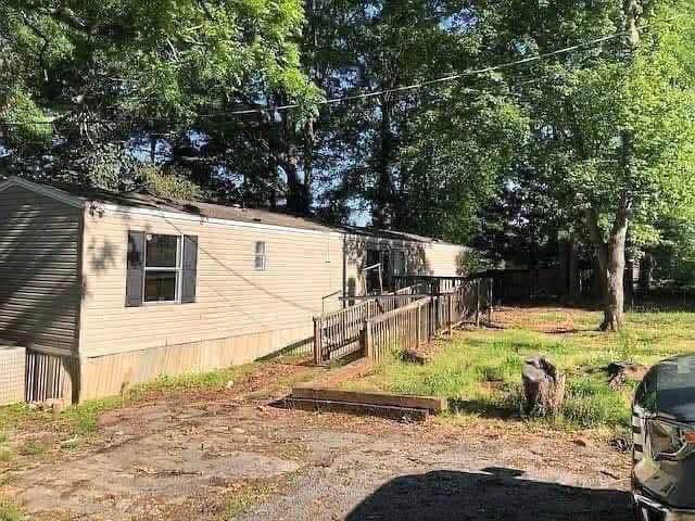 A beige mobile home with black shutters sits in a grassy yard surrounded by tall trees. A wooden ramp leads to the entrance, creating a serene, rural feel.