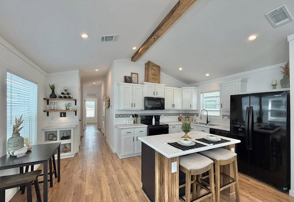 Modern kitchen with a vaulted ceiling and wooden beam features white cabinets, black appliances, and a central island with stools. Bright and airy feel.
