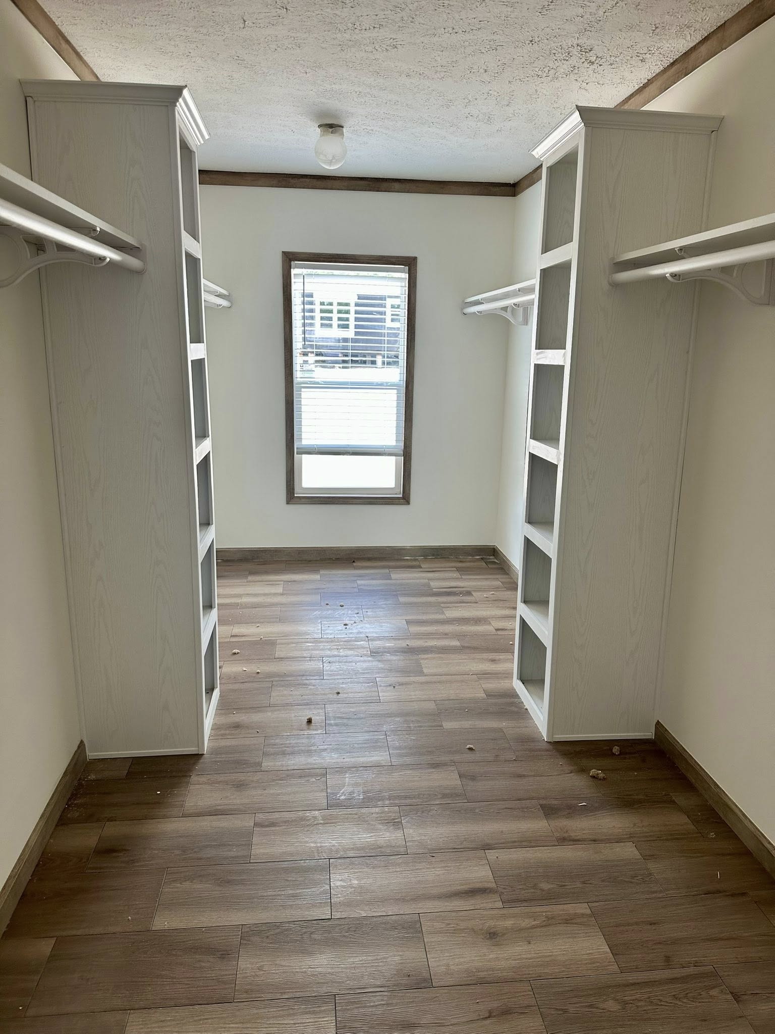 Spacious walk-in closet with white shelves and hanging rods on both sides, wooden floor, and a window at the far end letting in natural light.