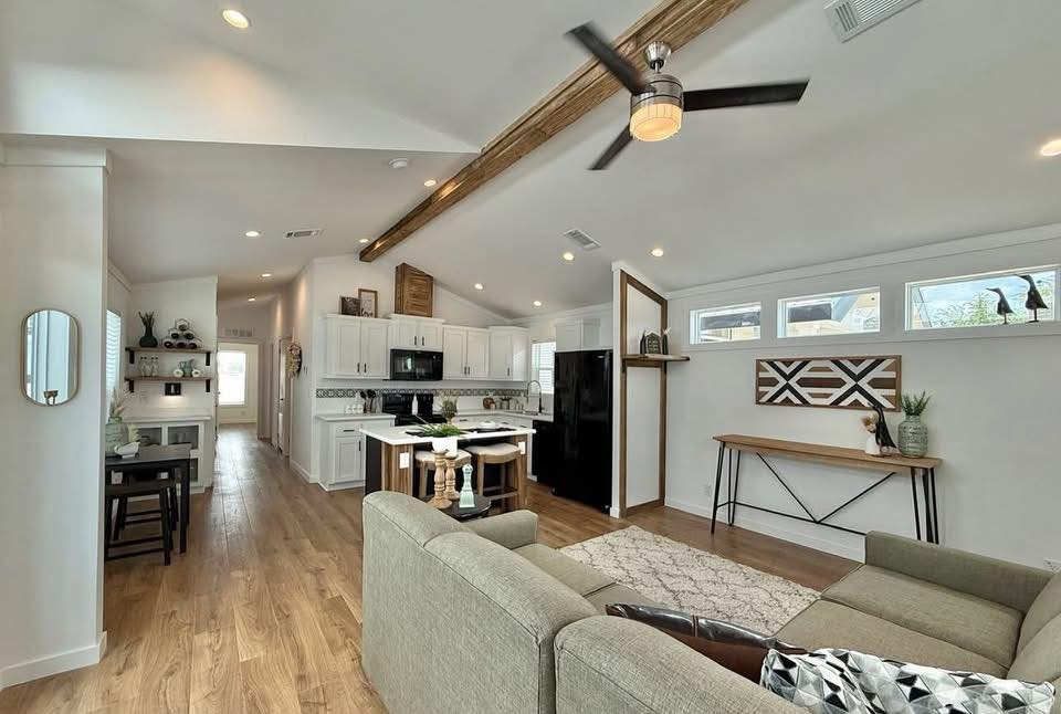 Open-concept living room and kitchen with vaulted ceilings, wooden beams, and hardwood floors. Modern decor features a gray sofa, geometric art, and plants.
