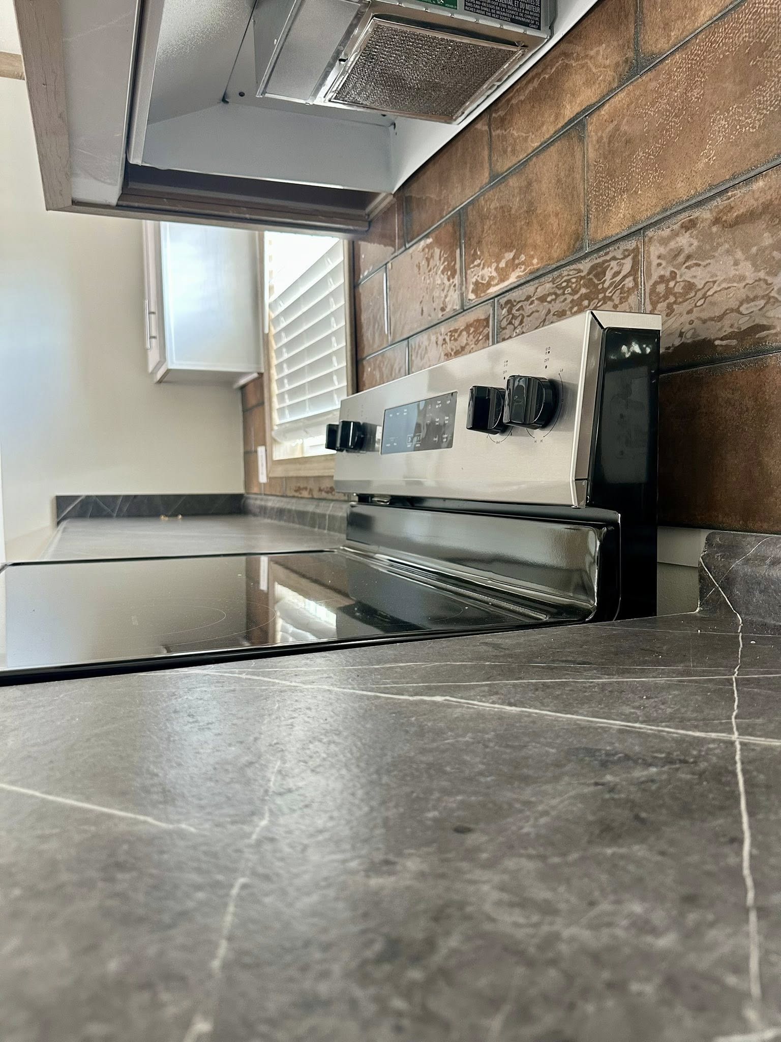 Modern kitchen with a shiny black stovetop and silver control panel, set against a warm brown tile backsplash. Sunlight filters through nearby blinds.