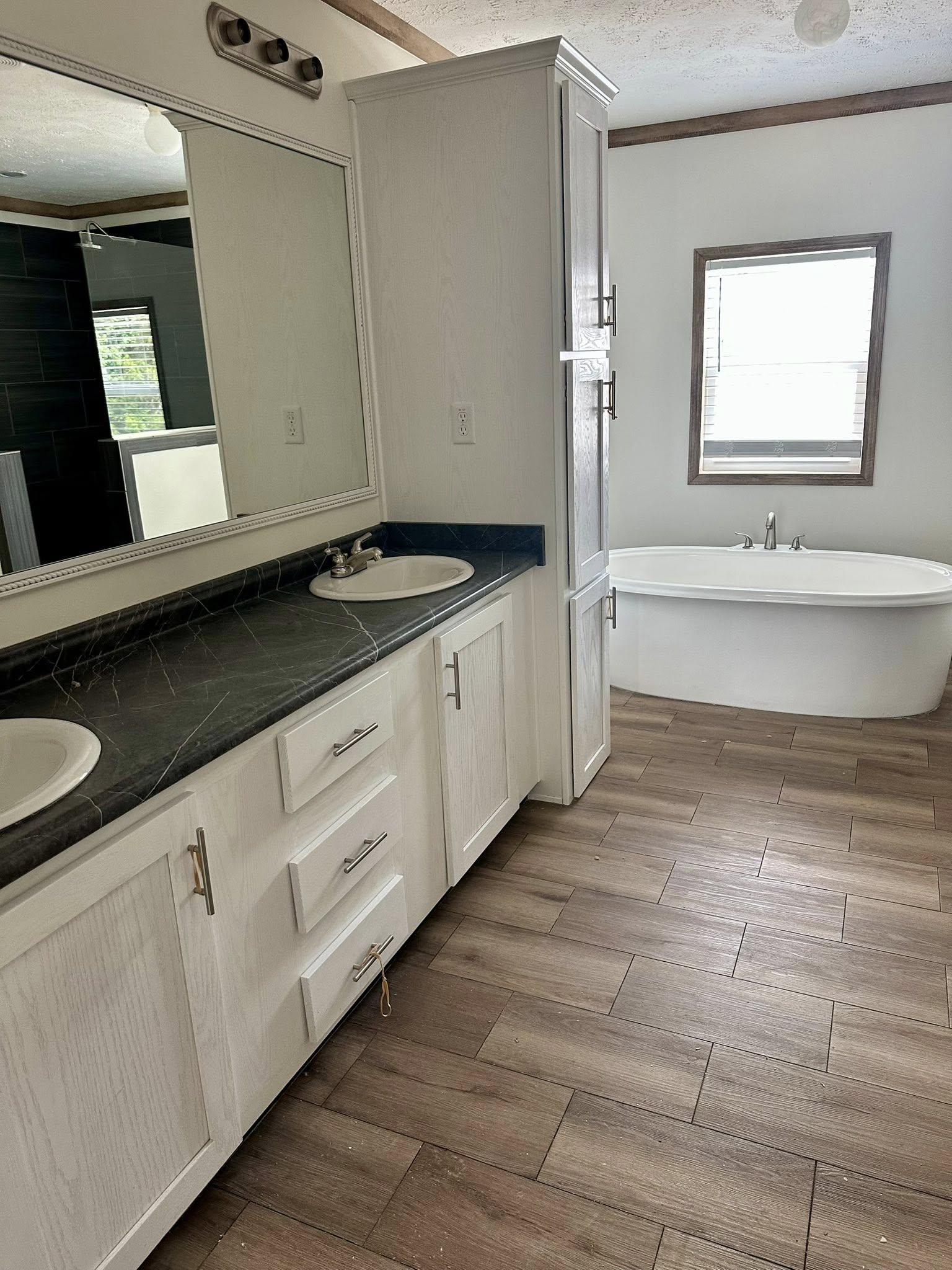 Spacious bathroom with double sinks on a dark countertop, a large mirror, and wood cabinets. A freestanding bathtub is seen near a window. Bright and elegant.