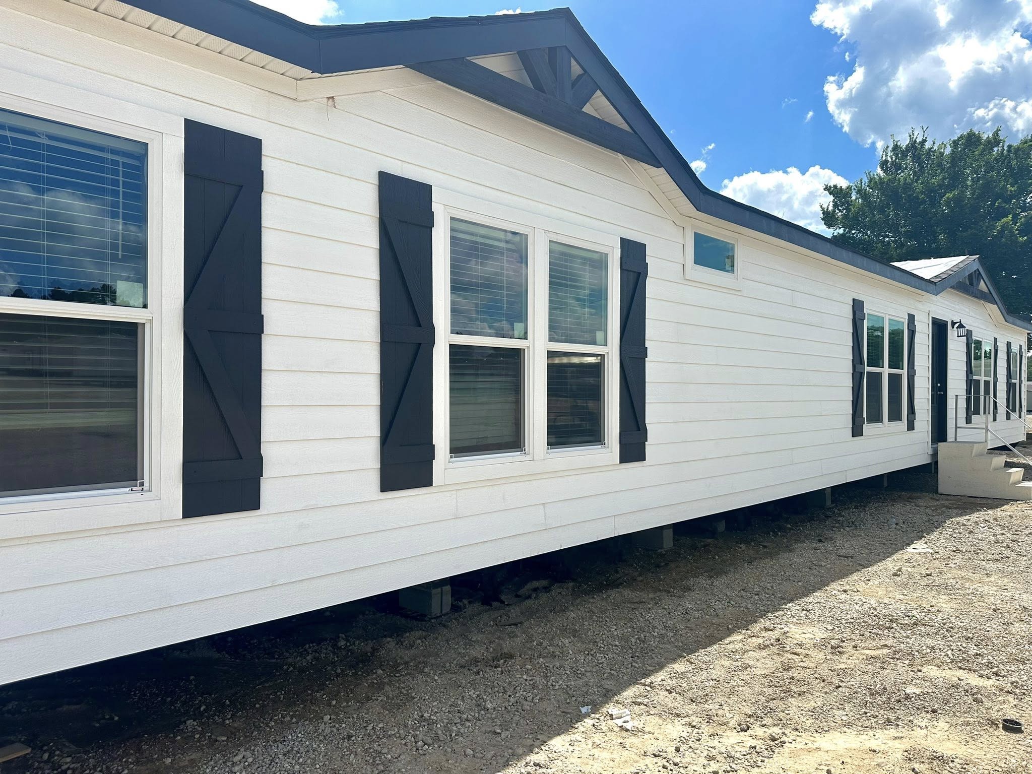 Long white mobile home with black shutters and windows, set on a gravel lot under a bright blue sky with clouds, surrounded by trees.
