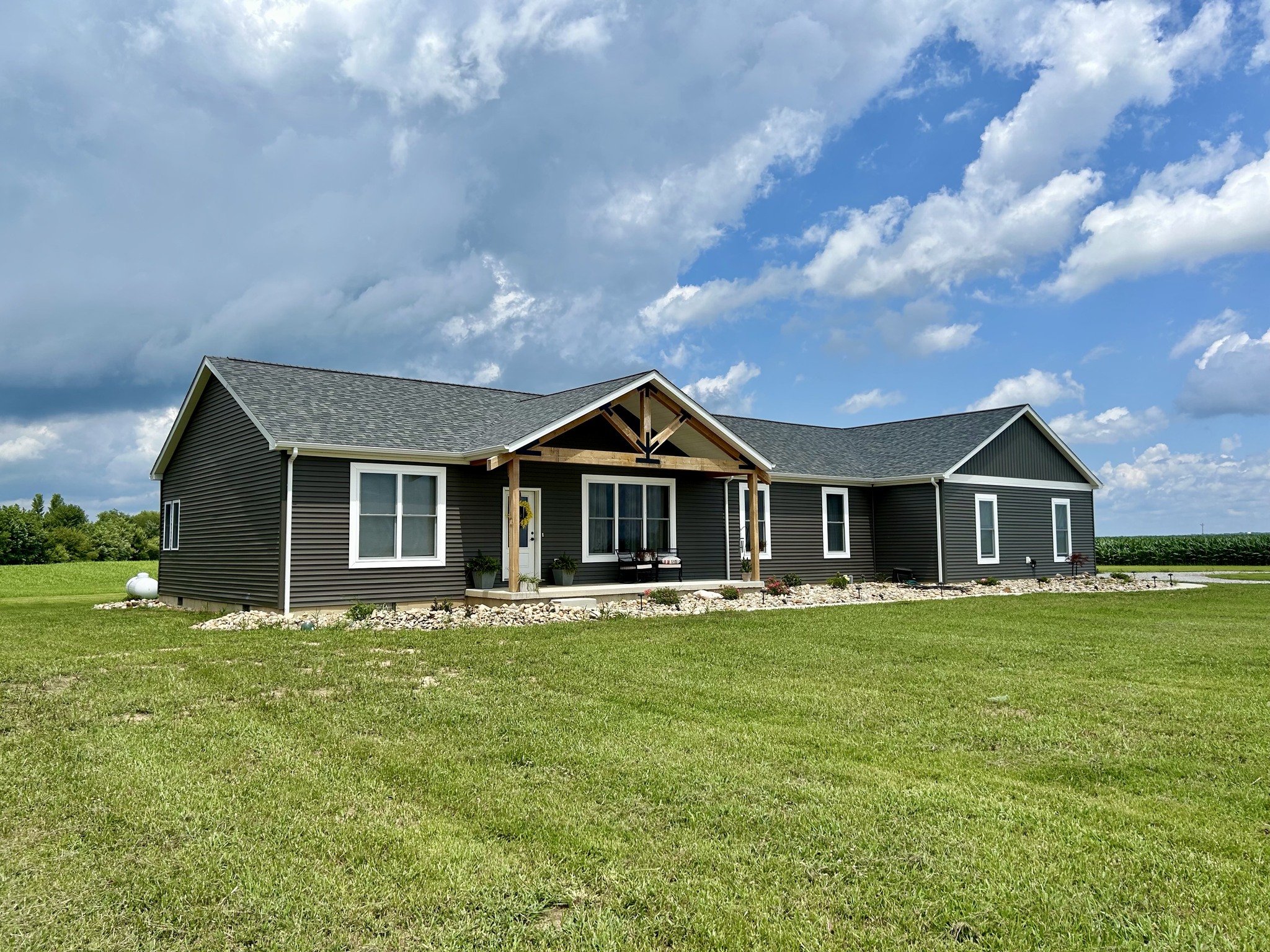 Single-story gray house with a wooden porch, surrounded by a lush green lawn under a partly cloudy blue sky. Calm, rural setting.