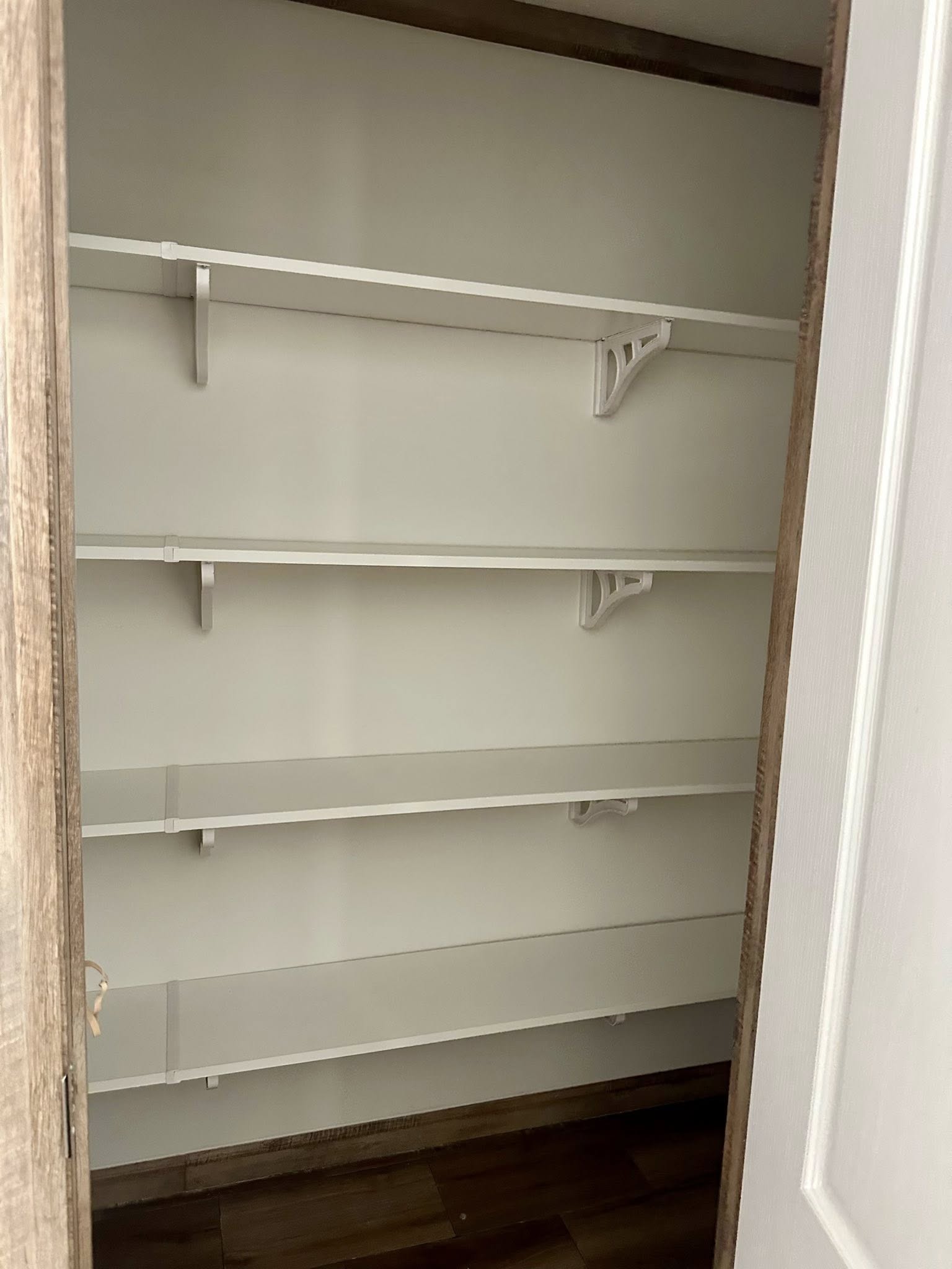 Empty closet with four white shelves, neatly installed with brackets against a light gray wall. Wooden floor and partial white door visible. Clean, orderly feel.