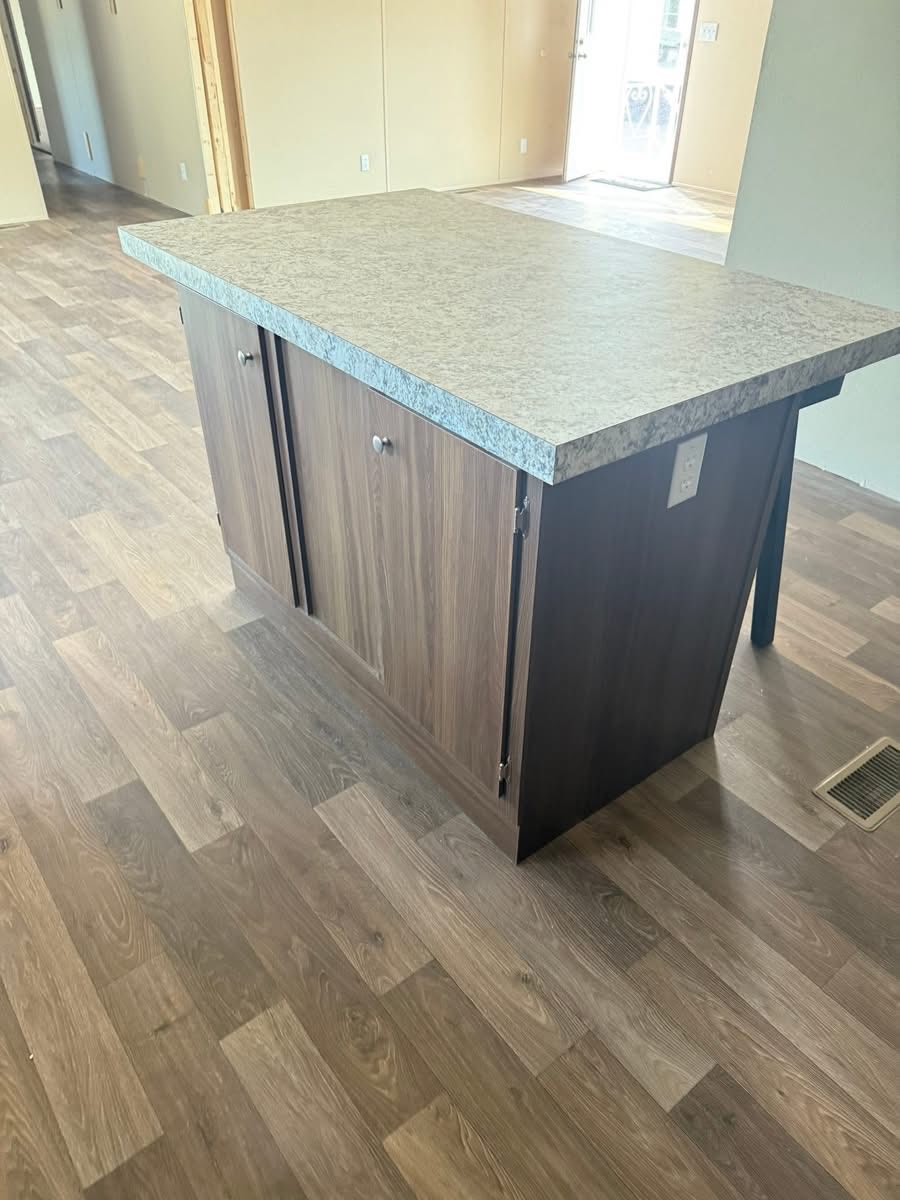 A wooden kitchen island with a speckled gray countertop stands on a light brown laminate floor. Natural light enters through a door in the background.