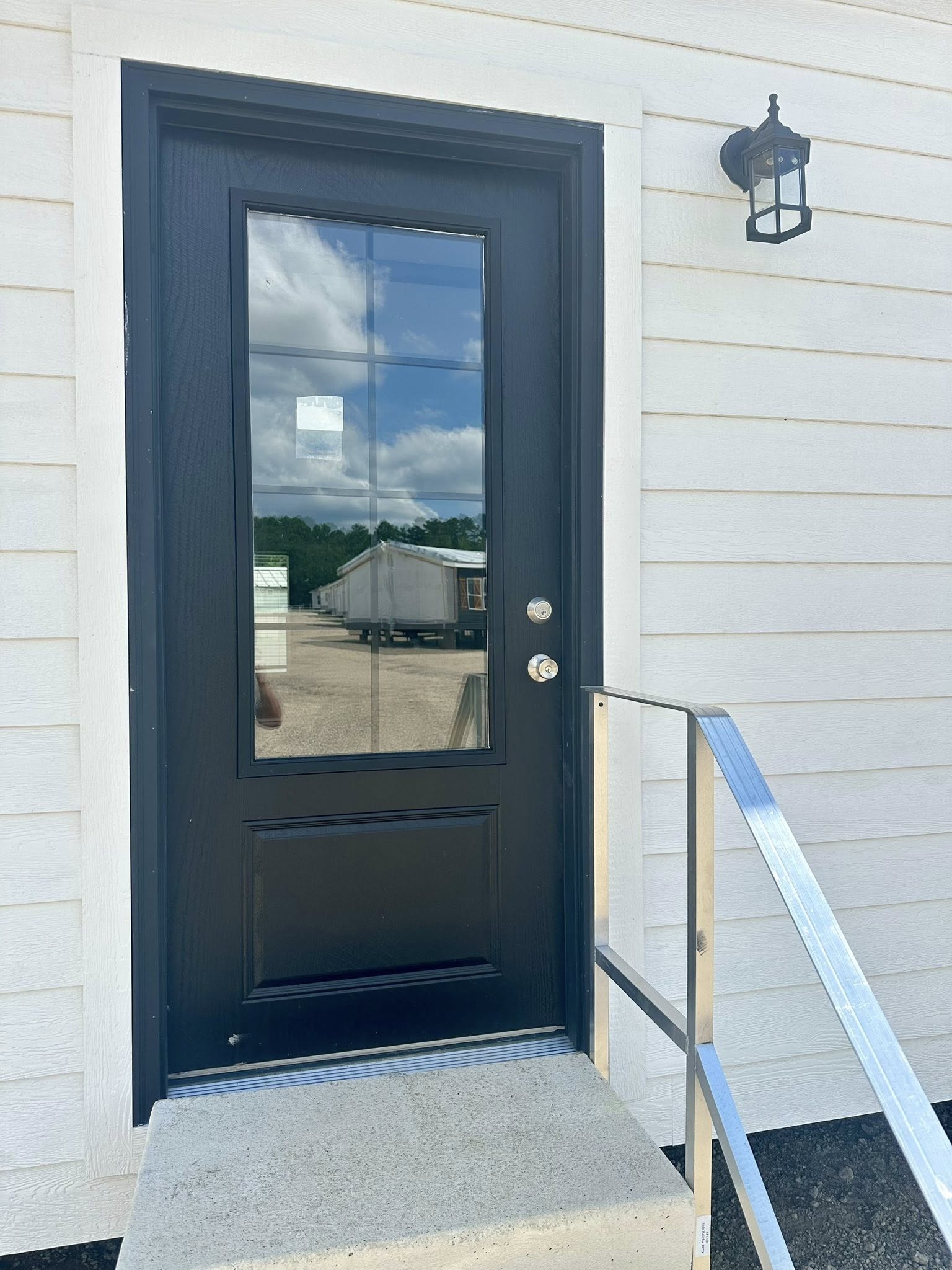Black door with glass panes reflecting a cloudy sky and trailer homes. Positioned on white siding, with a metal handrail and wall-mounted lamp.