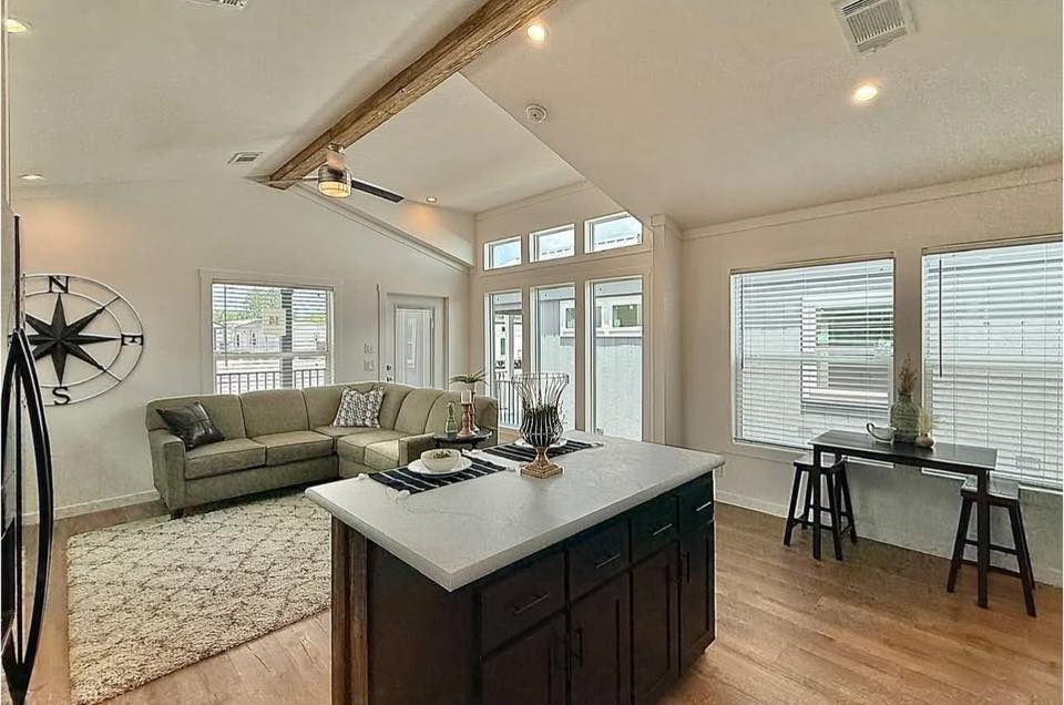 Spacious living area with modern decor, featuring a sectional sofa, a central island with dark cabinetry, large windows, and a decorative compass wall piece.