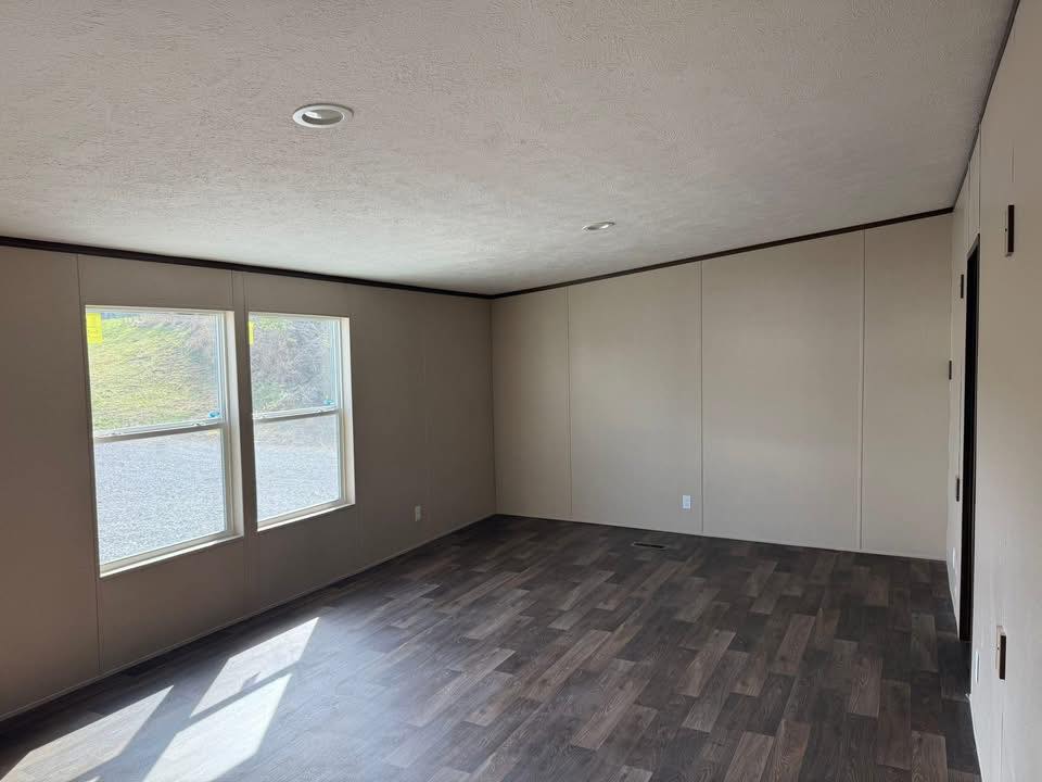 Empty room with beige walls and dark wood laminate flooring. Two large windows on the left let in natural light, creating a calm and inviting space.