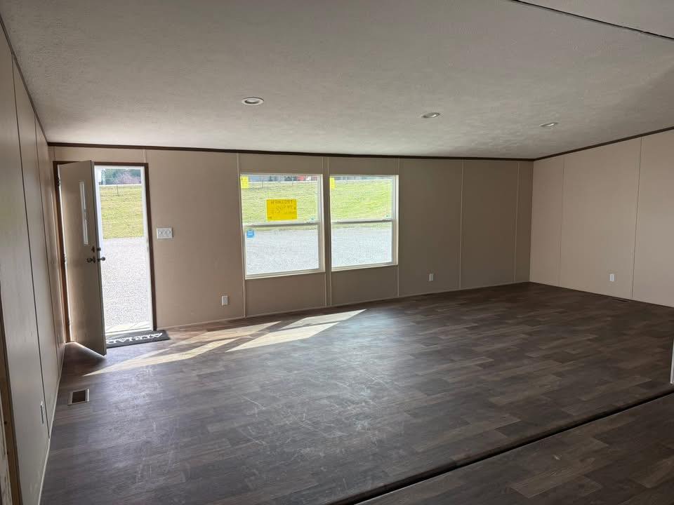 Empty room with dark hardwood floors, beige walls, and three large windows letting in natural light. The open door adds a sense of welcome and possibility.
