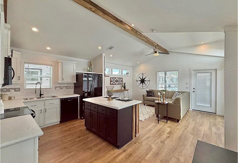 Bright, modern open-plan kitchen and living room. Features wood floors, white cabinets, dark island, sectional sofa, and decorative wall elements. Cozy and inviting space.