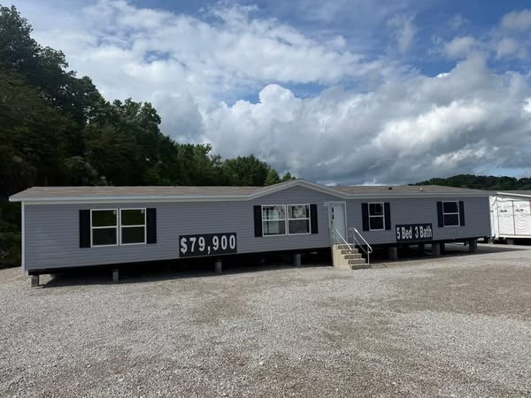 A gray mobile home on gravel, displaying "$79,900" and "5 Bed 3 Bath" in large letters. Cloudy sky and lush trees in the background convey a peaceful setting.
