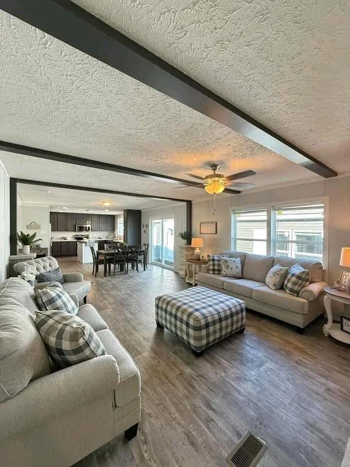 Spacious living room with gray sofas and plaid cushions, centered by a matching plaid ottoman. Open layout leads to a dining area and kitchen. Warm, cozy ambiance.