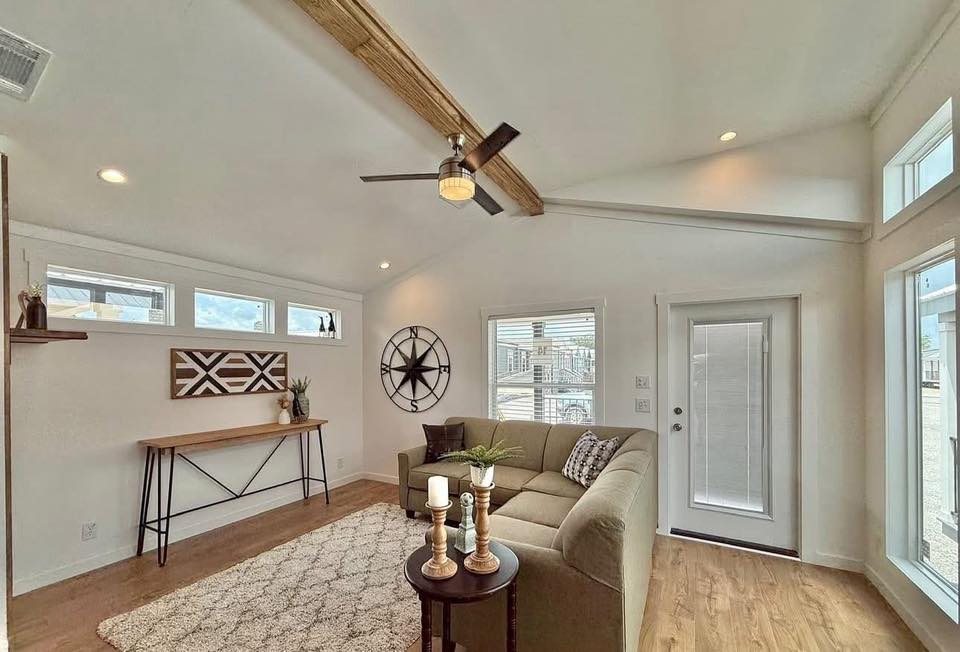 Bright living room with a vaulted ceiling, wooden beams, and a ceiling fan. Features a beige sectional sofa, decorative candles on a round table, abstract wall art, and a large clock. The room conveys a cozy, inviting atmosphere with ample natural light from the windows.