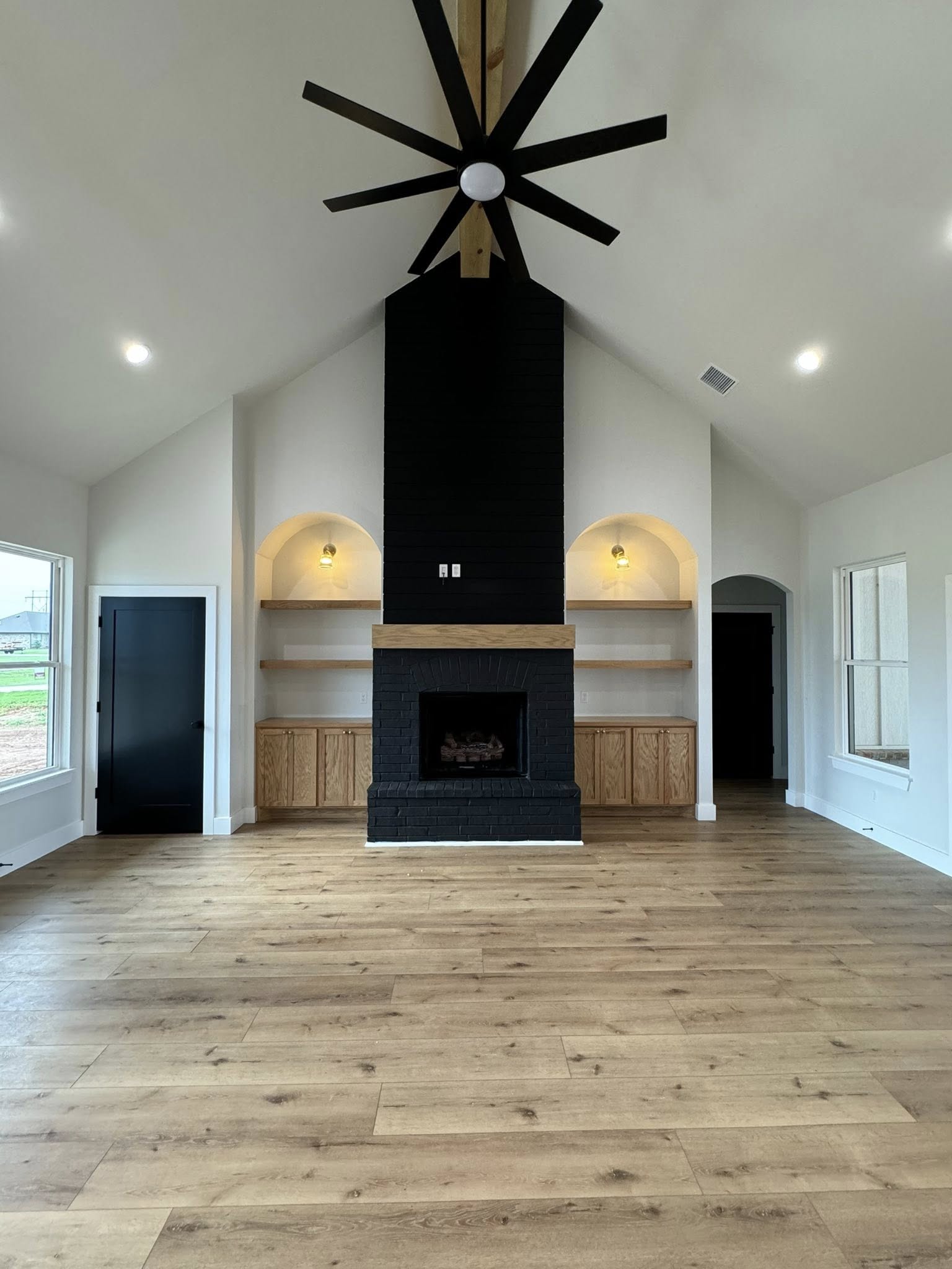 Spacious living room with vaulted ceiling, large black ceiling fan, black brick fireplace, arched shelves, light wood flooring, and bright atmosphere.