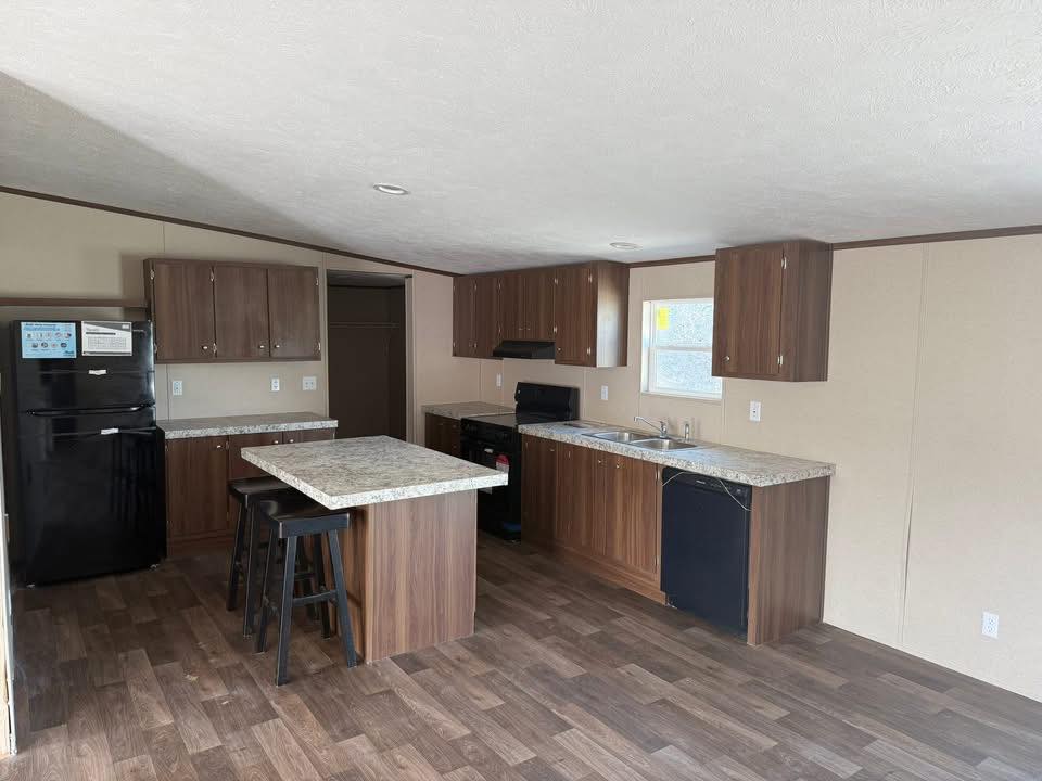 Modern kitchen with wood flooring, featuring dark wood cabinets, a large island with stools, black appliances, and a window providing natural light.