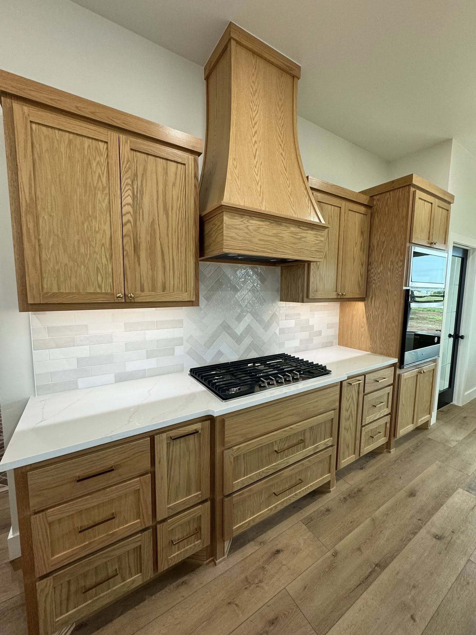 Modern kitchen with light wood cabinetry and sleek white countertops. Central gas stove sits beneath a decorative wood range hood. Warm, welcoming ambiance.