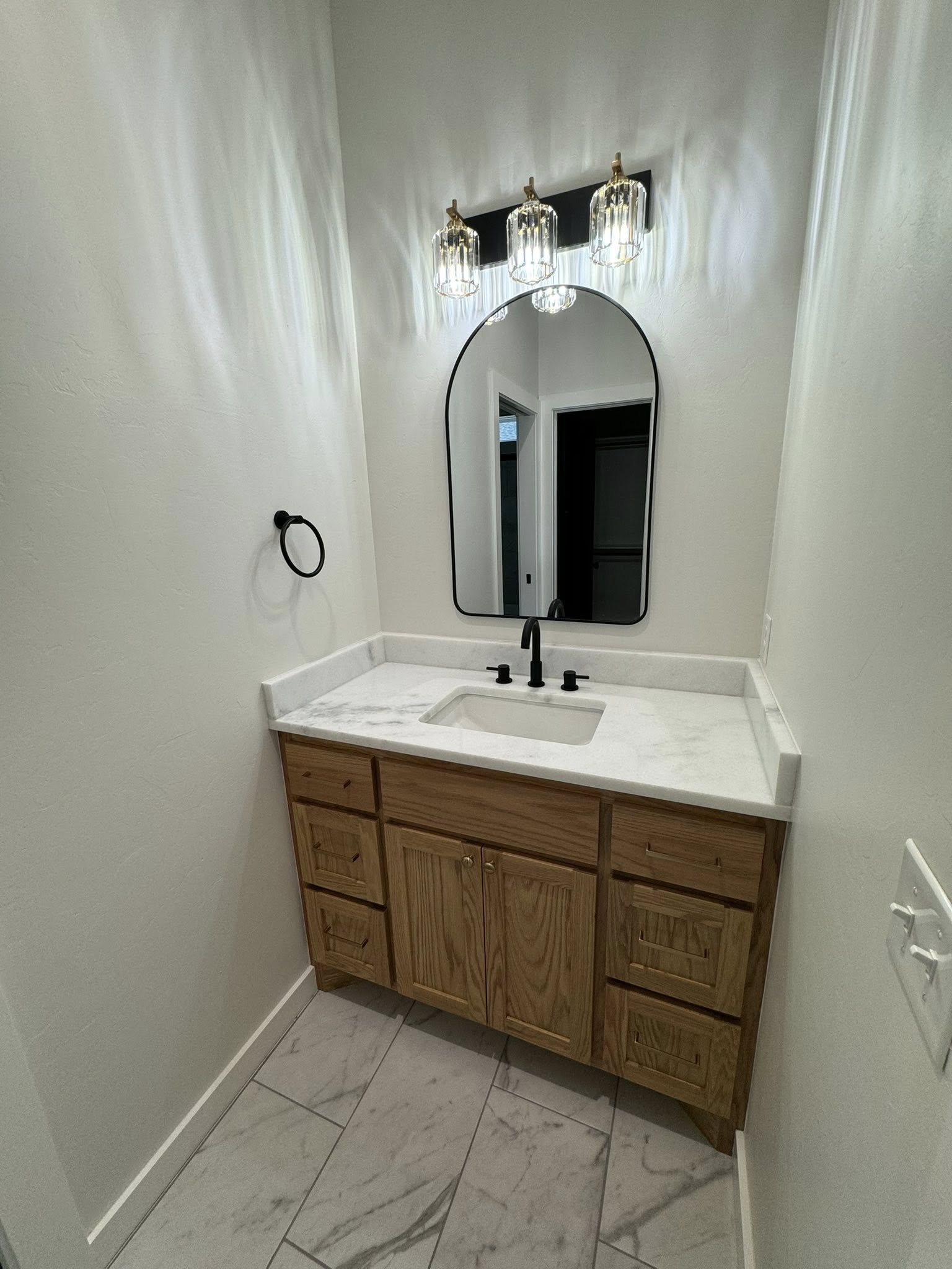 A small bathroom features a wooden vanity with a marble countertop and black faucet. An arched mirror is above, flanked by three elegant glass lights.