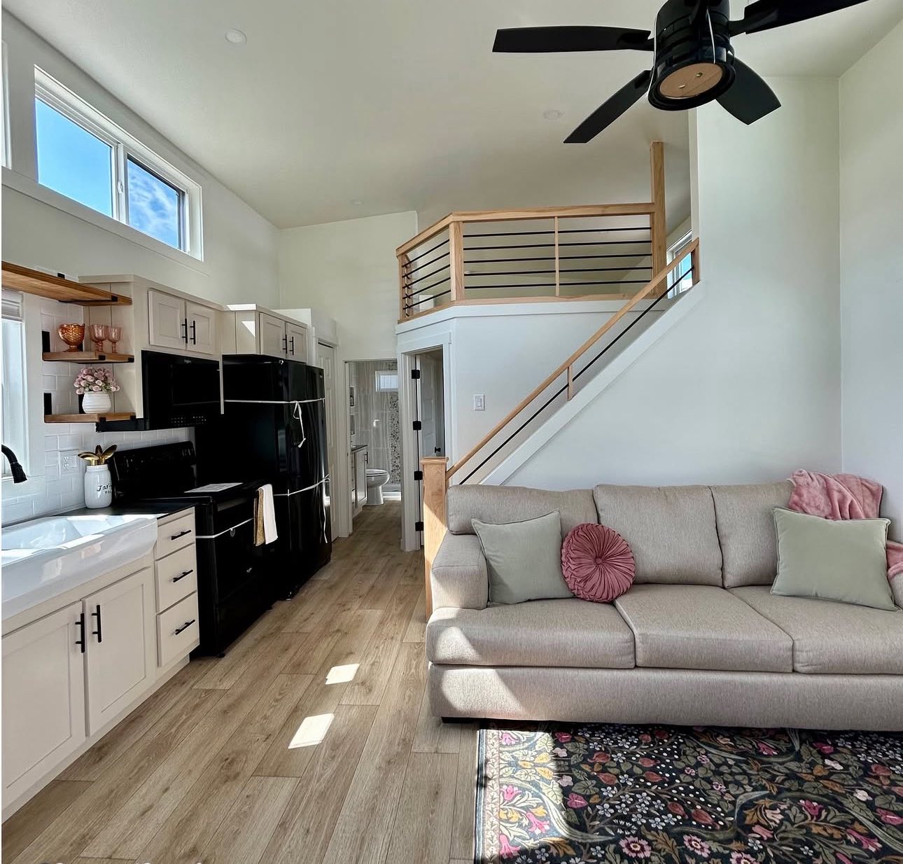 Modern tiny home interior with high ceiling and mezzanine. Beige couch with pink pillows, black appliances, white cabinets, and patterned rug.
