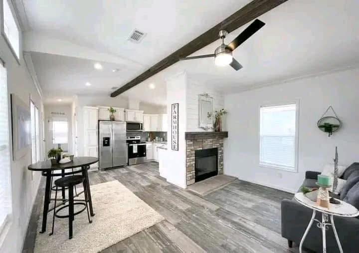 Modern open-concept living area with gray wood flooring, kitchen with stainless steel appliances, farmhouse decor, a cozy fireplace, and ceiling fan.