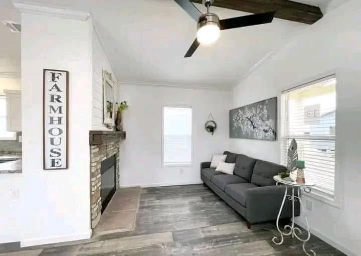 Bright living room with a farmhouse theme, featuring a stone fireplace, gray sofa, and white walls. Rustic decor includes a patterned rug and wall art. Ceiling fan overhead.