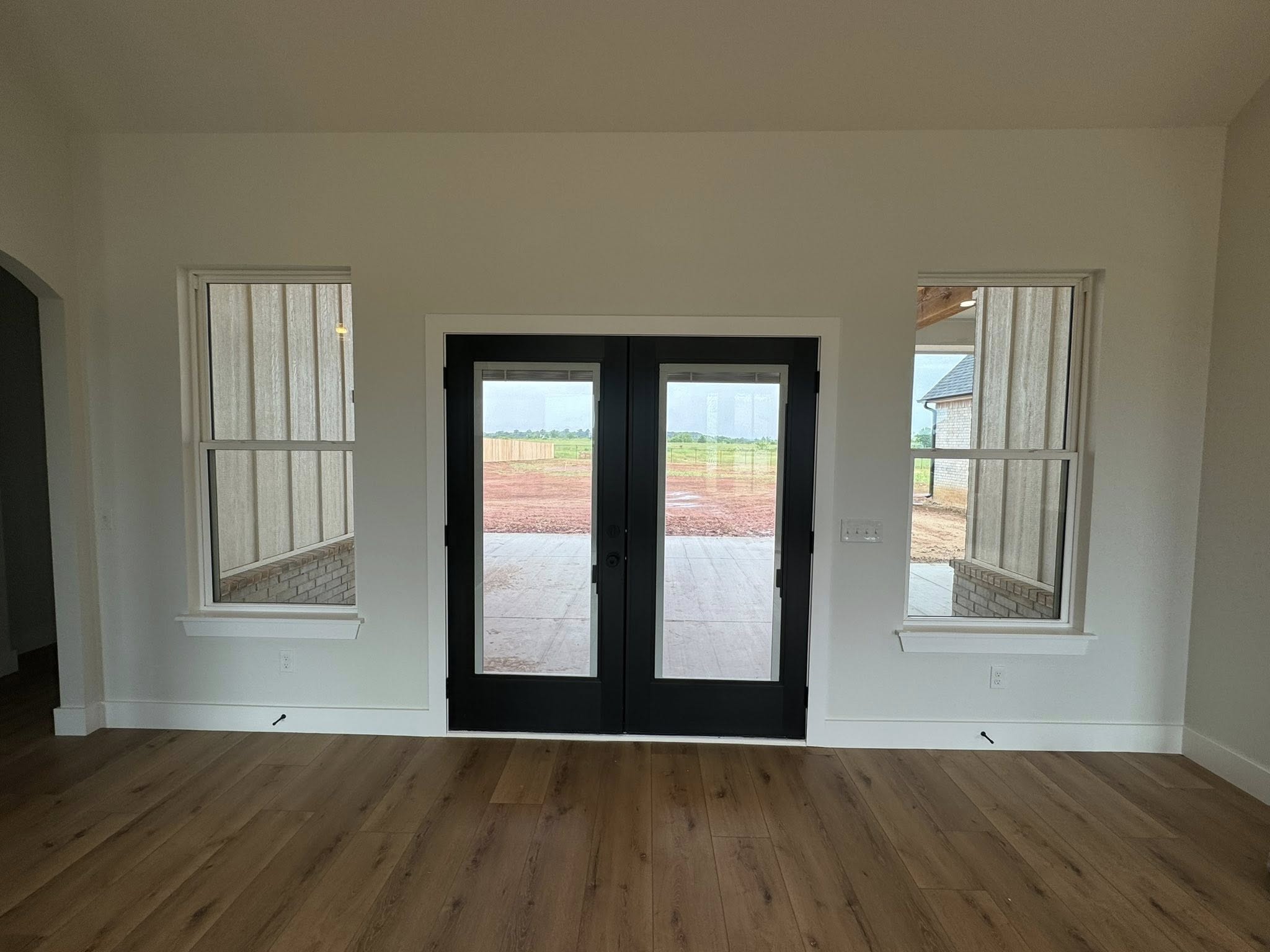 Interior room with light wood flooring, featuring black double doors flanked by two windows. The view outside shows a red dirt landscape.