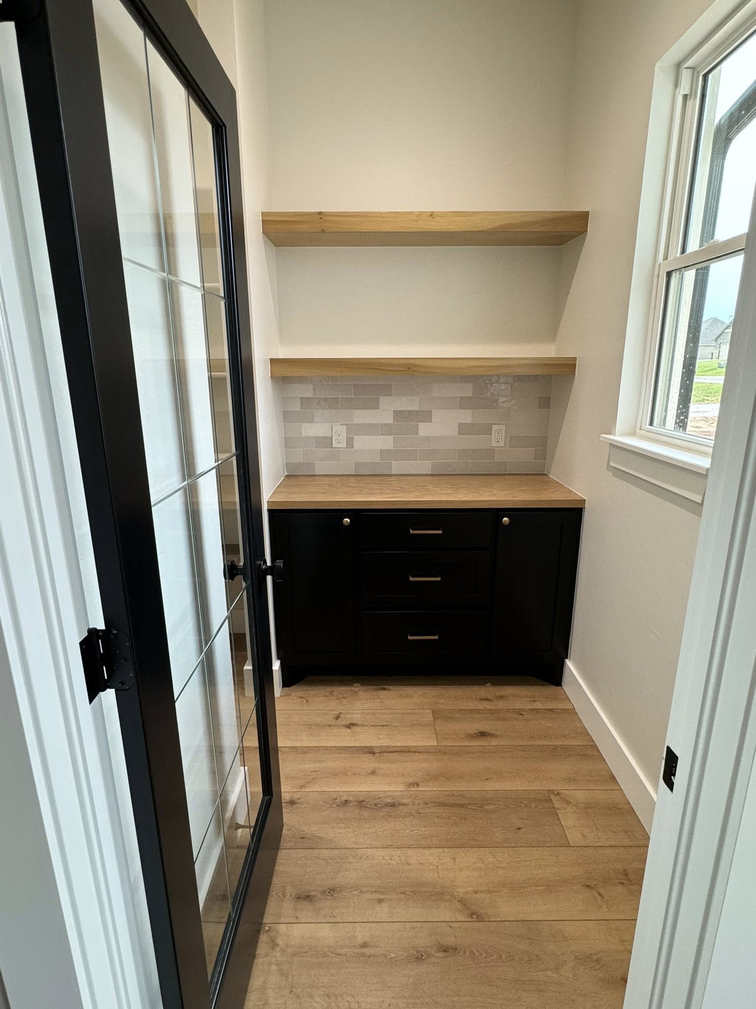 Narrow pantry with glass door, wood flooring, and two wooden shelves. Black cabinets with a wood countertop sit below a tiled backsplash. Bright ambiance.