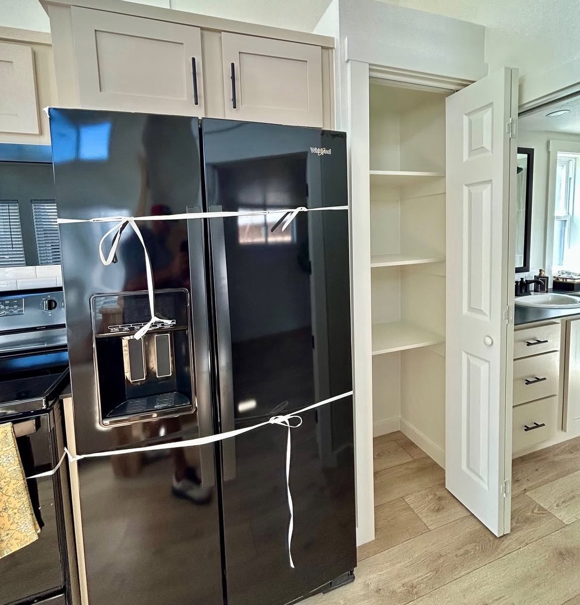 Black refrigerator secured with white rope, adjacent to an empty pantry with open white doors. Light wood floor, modern kitchen setting.