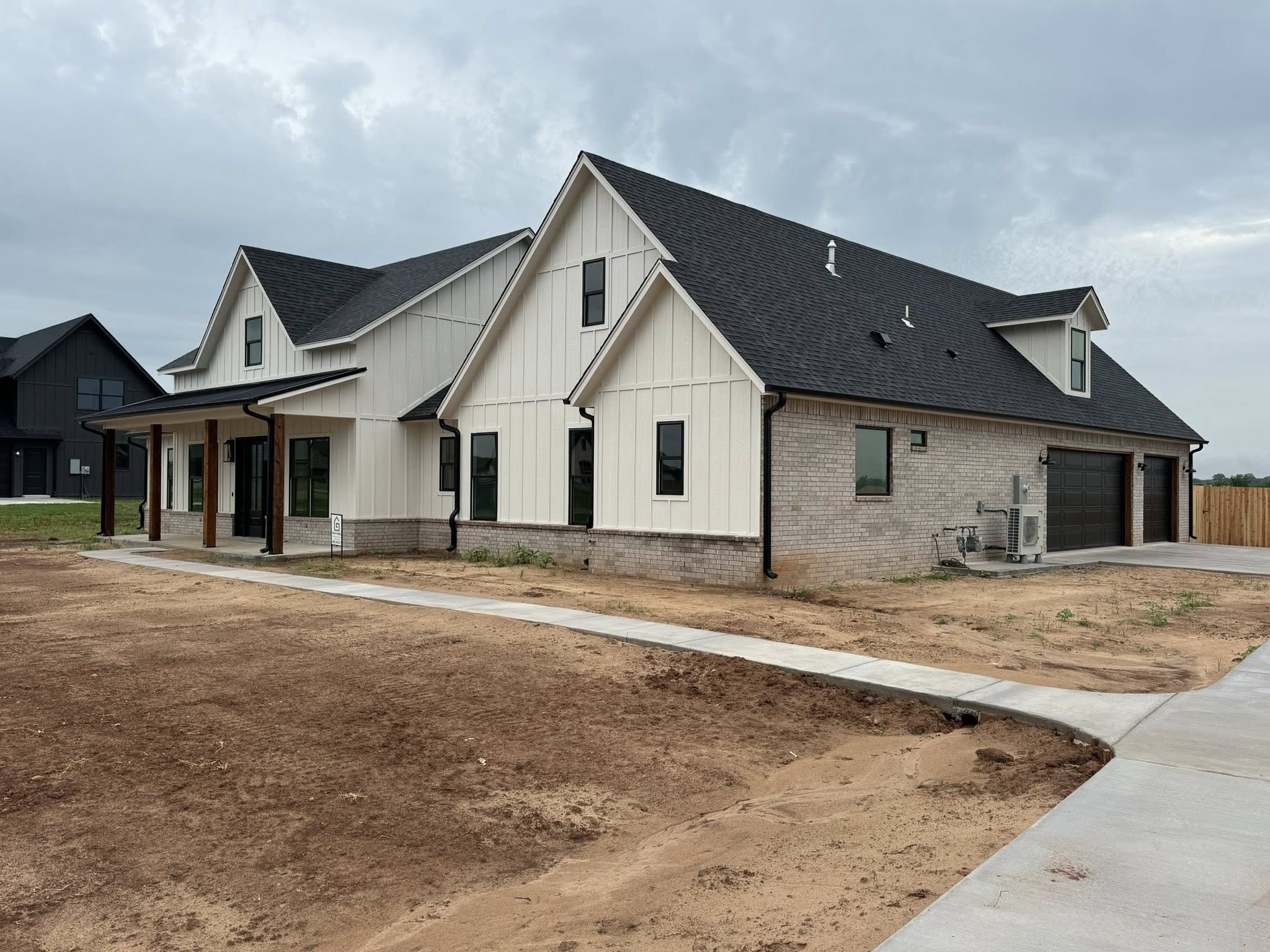 Modern farmhouse with white siding, brick accents, and a black roof. It sits on a barren yard under a cloudy sky, conveying a clean, rustic feel.