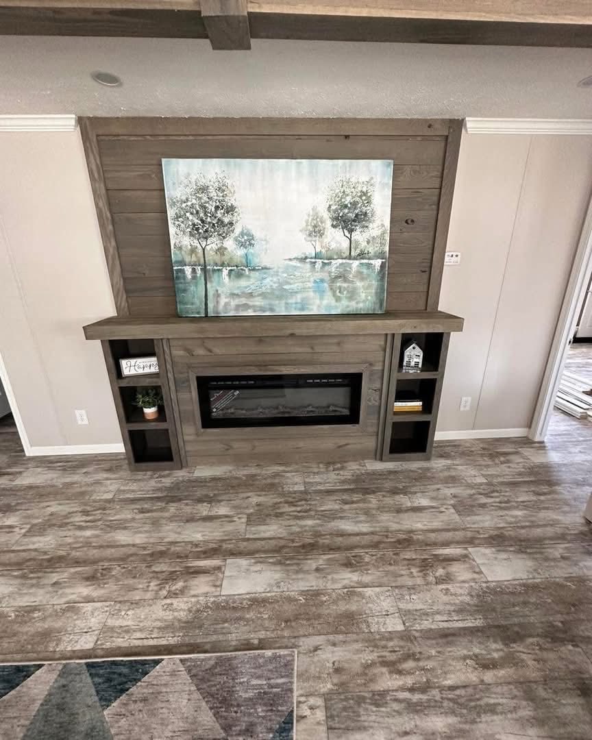 A cozy living room scene with a wood-paneled electric fireplace, topped by a serene landscape painting. Warm tones and decorative shelves complete the inviting ambiance.