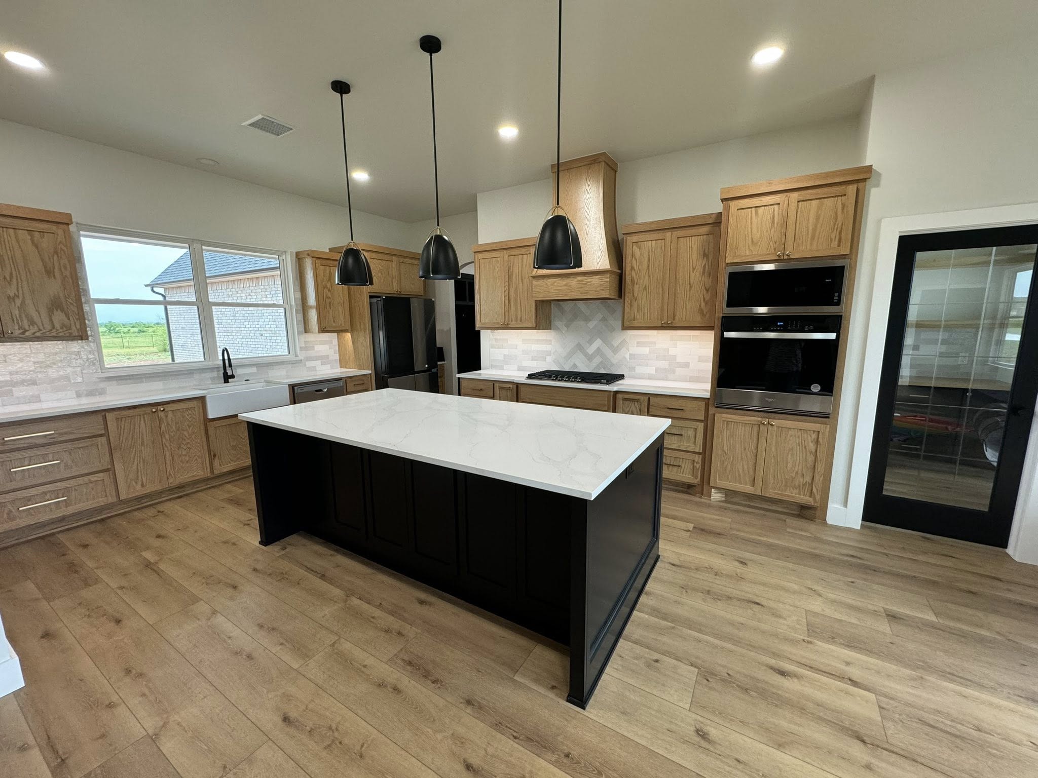 Modern kitchen with wood cabinets, white marble island, and black pendant lights. Natural light from a large window and a glass door; hardwood floors.