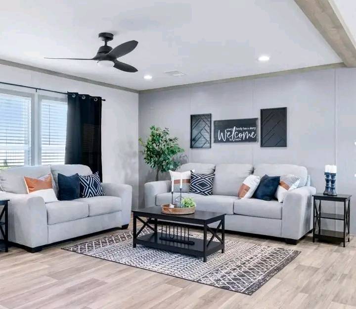 A bright living room with two gray sofas, patterned cushions, a black ceiling fan, a coffee table, and wall art saying "Welcome," creating a cozy atmosphere.