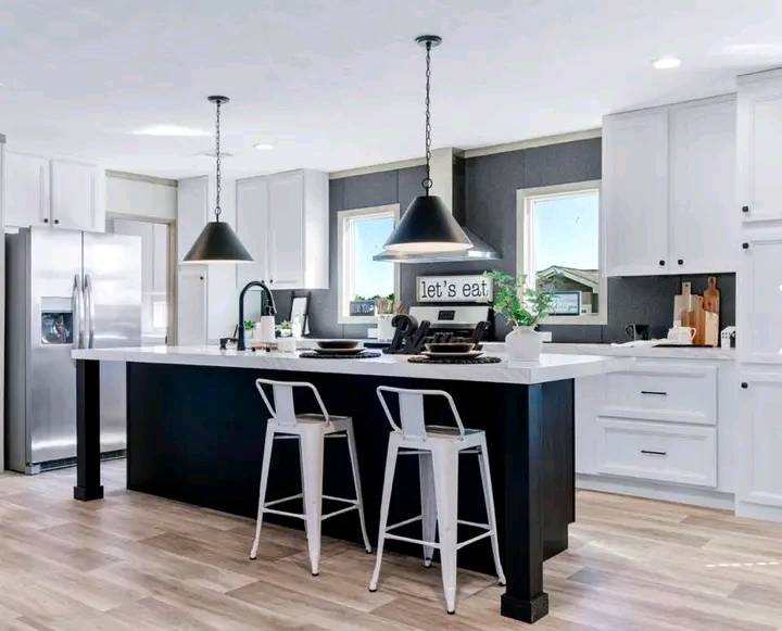 Modern kitchen with white cabinets, stainless steel appliances, and a black island. Two pendant lights hang above the island with two white stools. A sign reads "let's eat."