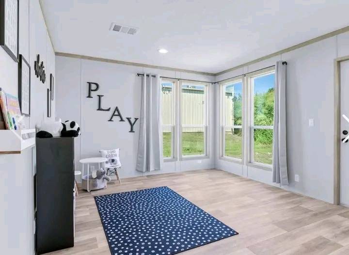 Bright playroom with wooden floors, a polka dot rug, and large windows with gray curtains. "PLAY" letters are mounted on the wall, creating a cheerful atmosphere.