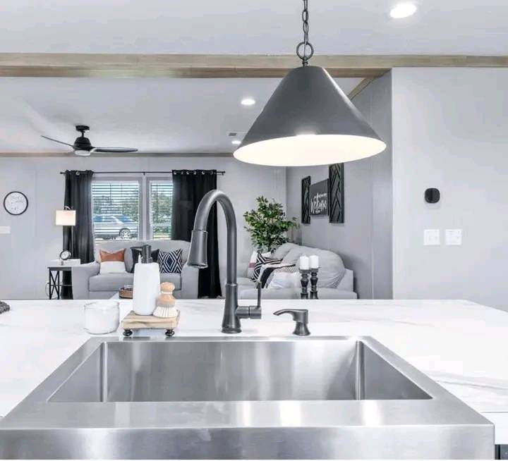 Modern kitchen with a stainless steel sink in the foreground, black pendant light above. Sleek living area with gray sofa, black curtains, and decor. Cozy ambiance.