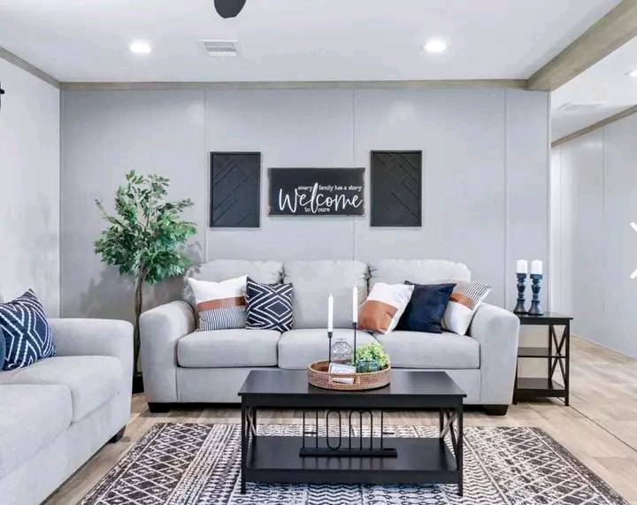 A cozy living room with a gray sofa adorned with mixed cushions and a "Welcome" sign on the wall. A coffee table holds candles and a plant, creating a warm atmosphere.