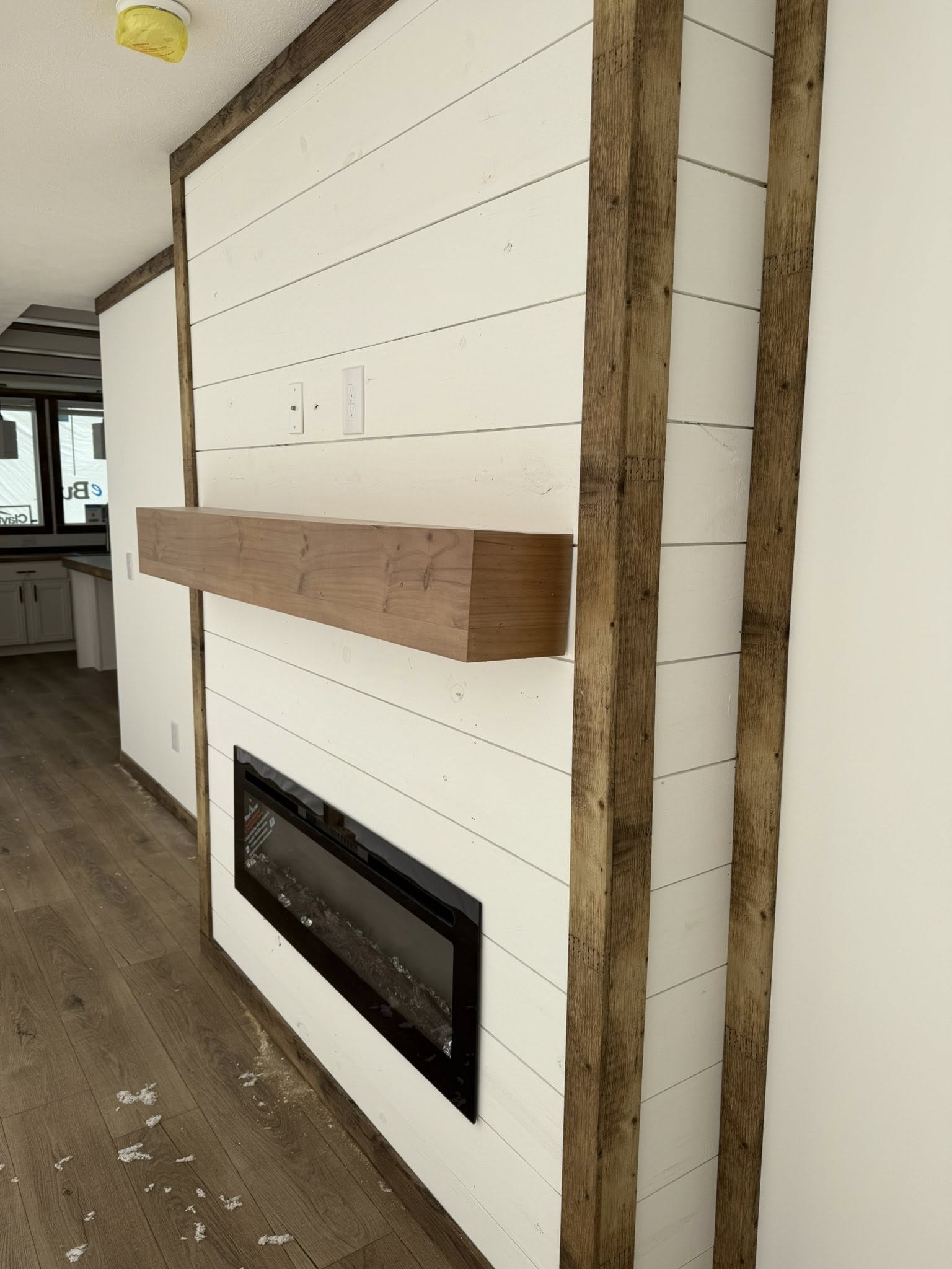 White shiplap wall with dark wood trim features a sleek black fireplace insert. A wooden mantel is above, conveying a modern rustic style.