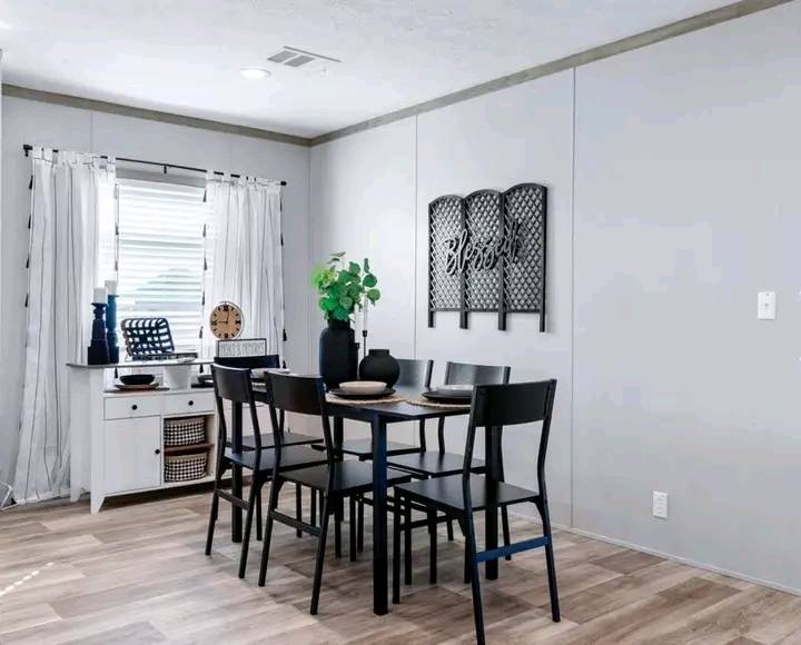 A modern dining room with a sleek black table and six chairs, set for dining. A white cabinet with decor is near a window with sheer curtains. A wall art with the word "Blessed" adds warmth.